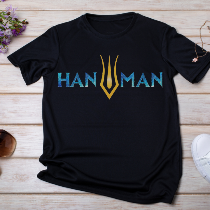 Hanuman-themed t-shirt with bold typography design featuring a golden trident logo in the center, displayed flat on a wooden background.